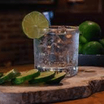 Ein Glas mit klarem Getränk, Eis und einem Limettenrad steht auf einem Holzbrett neben einer in Scheiben geschnittenen Gurke - die perfekte Erfrischung für Veranstaltungen. Im Hintergrund sorgt eine Schale mit ganzen Limetten für eine lebendige Note.