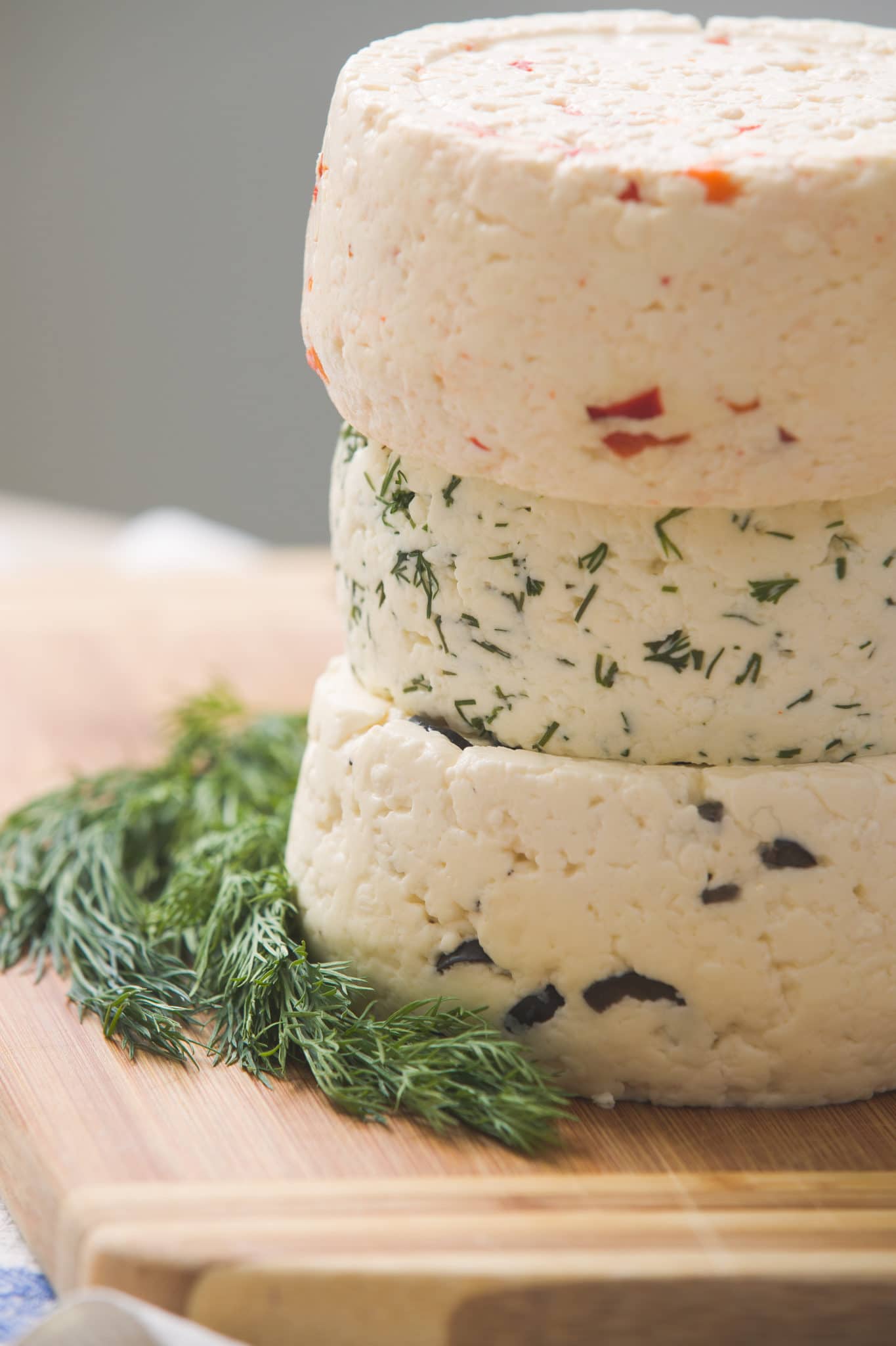 Auf einem hölzernen Schneidebrett sind drei Runden hausgemachter Käse mit verschiedenen Kräutern und Gewürzen gestapelt - die perfekte Inspiration für Profis oder Besucher von Fachseminaren. Frischer Dill wird neben den Käse gelegt.