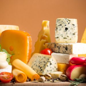 Eine Vielzahl von Käsesorten - darunter gelbe, blau geäderte und mit Rinde überzogene Sorten - sind kunstvoll mit Kirschtomaten, Kräutern und Nüssen auf einer Holzfläche arrangiert und bilden vor einem warmen orangefarbenen Hintergrund eine Auslage, die eines Profis oder Fachseminares würdig ist.