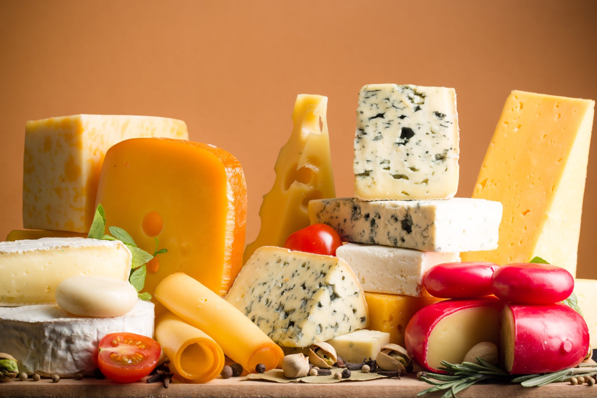 Eine Vielzahl von Käsesorten - darunter gelbe, blau geäderte und mit Rinde überzogene Sorten - sind kunstvoll mit Kirschtomaten, Kräutern und Nüssen auf einer Holzfläche arrangiert und bilden vor einem warmen orangefarbenen Hintergrund eine Auslage, die eines Profis oder Fachseminares würdig ist.