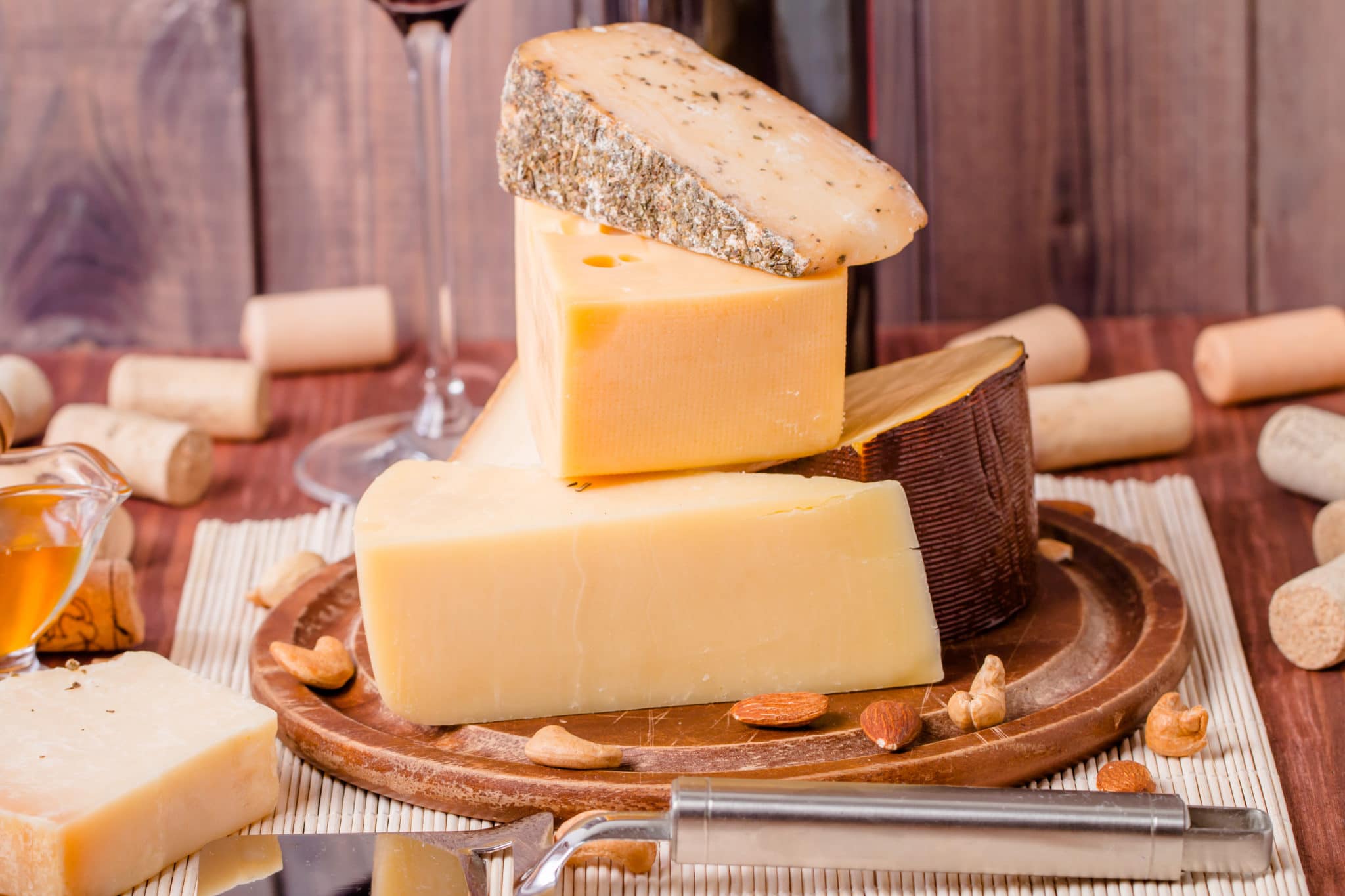 Ein Holzbrett mit gestapelten Käseblöcken, umgeben von Nüssen, einem Käsemesser, Honig, Weingläsern und Weinkorken auf einem rustikalen Tisch - perfekt für Profis oder Fachseminare in Gourmet-Degustationen.