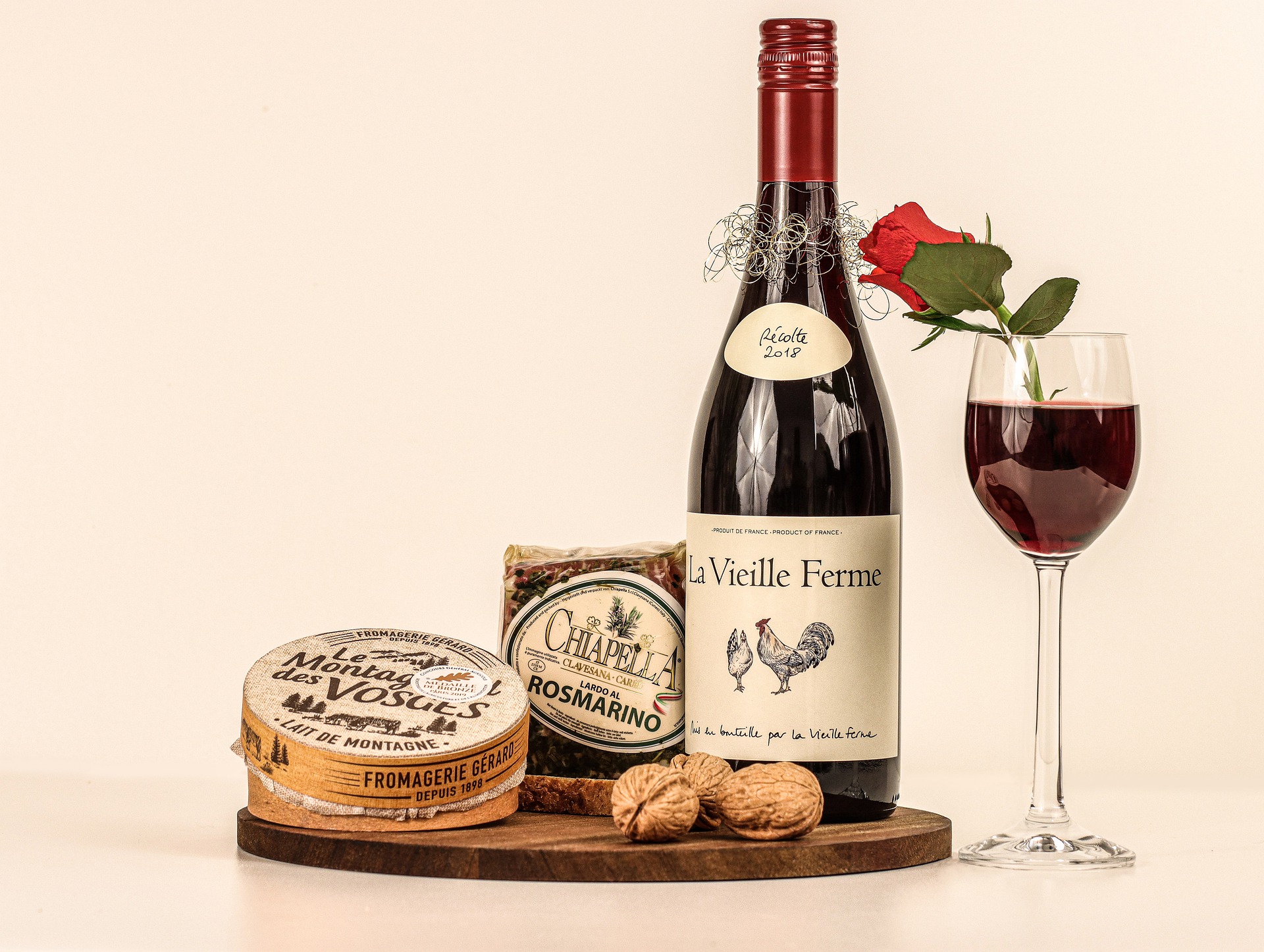 Eine Flasche Rotwein, ein Glas mit einer roten Rose, Käse, Rosmarin-Cracker und Walnüsse sind elegant auf einem Holzbrett arrangiert - perfekt für Veranstaltungen oder intime Zusammenkünfte vor einem schlichten Hintergrund.