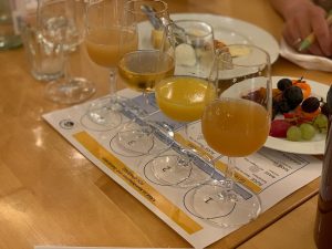 Auf einem Holztisch stehen vier Gläser mit trübem Apfelwein auf einer Probiermatte, daneben ein Teller mit Käse und Brot und ein weiterer Teller mit Weintrauben und Obst. Im Hintergrund ist eine Hand zu sehen, die einen grünen Marker hält.