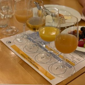 Auf einem Holztisch stehen vier Gläser mit trübem Apfelwein auf einer Probiermatte, daneben ein Teller mit Käse und Brot und ein weiterer Teller mit Weintrauben und Obst. Im Hintergrund ist eine Hand zu sehen, die einen grünen Marker hält.