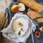 Ein runder, auf Pergament ausgeschnittener Käse, belegt mit zwei grünen Oliven, bildet den Rahmen für eine köstliche Degustation - auf einem grauen Tuch platziert, mit Messer, Baguettescheiben, Feigen, Äpfeln, Birnen und Weizenstängeln in der Nähe. Vive la France im Jahr 2025!.