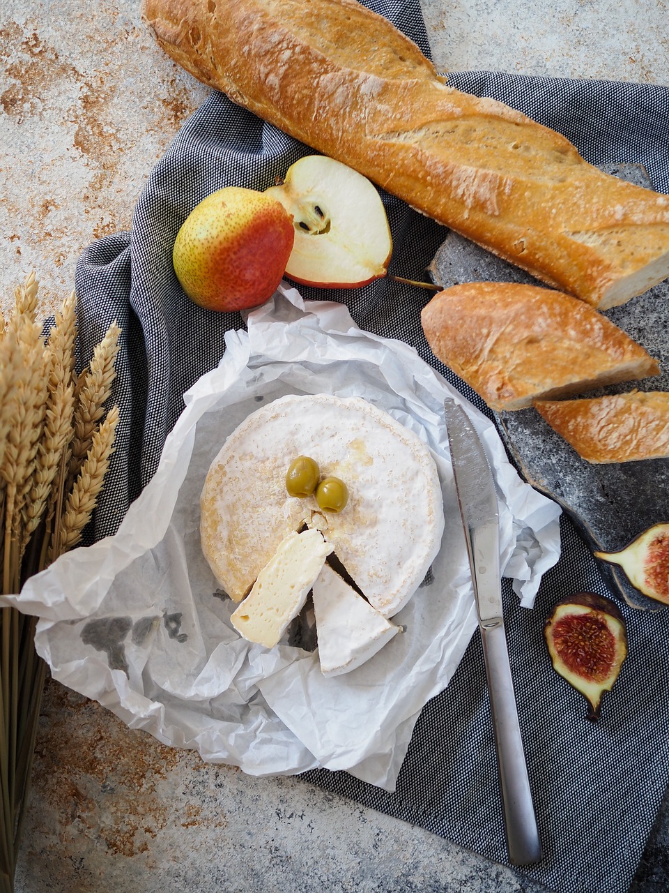 Ein runder, auf Pergament ausgeschnittener Käse, belegt mit zwei grünen Oliven, bildet den Rahmen für eine köstliche Degustation - auf einem grauen Tuch platziert, mit Messer, Baguettescheiben, Feigen, Äpfeln, Birnen und Weizenstängeln in der Nähe. Vive la France im Jahr 2025!.