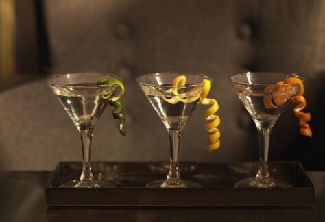 Auf einem Tablett stehen drei Martini-Gläser mit klaren Wermut-Cocktails, jedes garniert mit einer gewellten Zitrusschale - grün, gelb und orange - vor einem verschwommenen, dunklen Hintergrund.