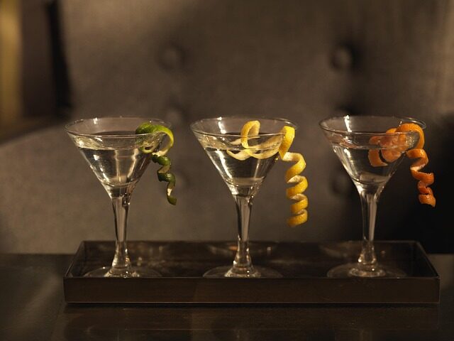 Auf einem Tablett stehen drei Martini-Gläser mit klaren Wermut-Cocktails, jedes garniert mit einer gewellten Zitrusschale - grün, gelb und orange - vor einem verschwommenen, dunklen Hintergrund.