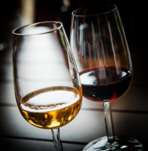 Zwei Weingläser stehen nebeneinander auf einem Tisch, eines mit Weißwein, das andere mit Rotwein. Die Gläser reflektieren das Licht und heben die Farben und die Klarheit der Weine hervor - die perfekte Ergänzung zu einem Käsebuffet.