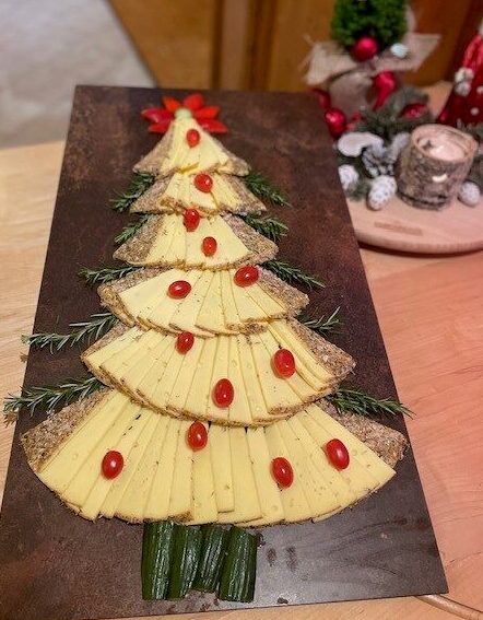 Eine festliche Käseplatte in Form eines Weihnachtsbaums, mit Schichten von Käsescheiben und Brot, Traubentomaten als Ornamente, Rosmarinzweigen als Äste und grünem Gemüse als Baumstamm.