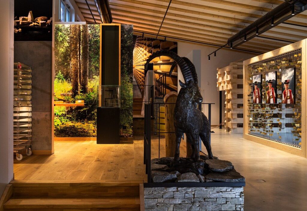 Ein modernes Museumsinterieur mit Holzbalken, einem Steinsockel, auf dem eine lebensgro&szlig;e Steinbockstatue steht, lebhafter Waldkunst, Vitrinen und historischen Fotos an den W&auml;nden. Warme Beleuchtung unterstreicht die Exponate.