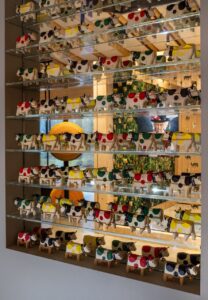 Ein Regal mit mehreren Reihen kleiner, dekorativer Kuhfiguren, die jeweils mit verschiedenen Farbflecken bemalt sind, steht vor einem Spiegel, der einen modernen, warm beleuchteten Raum mit Pflanzen und einer runden orangefarbenen Lampe widerspiegelt.