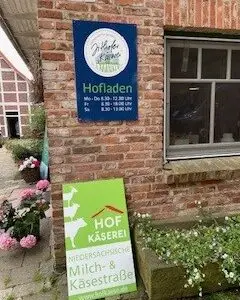 Ein Backsteingebäude mit zwei Schildern: ein blaues für den Hofladen mit den Öffnungszeiten und ein grünes für die Hof Käserei an der Niedersächsischen Milch- & Käsestraße. In der Nähe befinden sich rosa Blumen.
