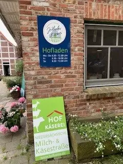 Ein Backsteingebäude mit zwei Schildern: ein blaues für den Hofladen mit den Öffnungszeiten und ein grünes für die Hof Käserei an der Niedersächsischen Milch- & Käsestraße. In der Nähe befinden sich rosa Blumen.