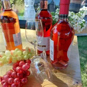 Drei Flaschen Roséwein aus dem Weinland, zwei Weingläser, dekorative grüne und rote Weintrauben und weiße Blumen auf einem Holztisch im Freien - perfekt für Ihre Think Pink Feier am 18.09.2026 mit Grünzeug und Bannern im Hintergrund.