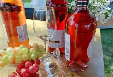 Drei Flaschen Roséwein aus dem Weinland, zwei Weingläser, dekorative grüne und rote Weintrauben und weiße Blumen auf einem Holztisch im Freien - perfekt für Ihre Think Pink Feier am 18.09.2026 mit Grünzeug und Bannern im Hintergrund.