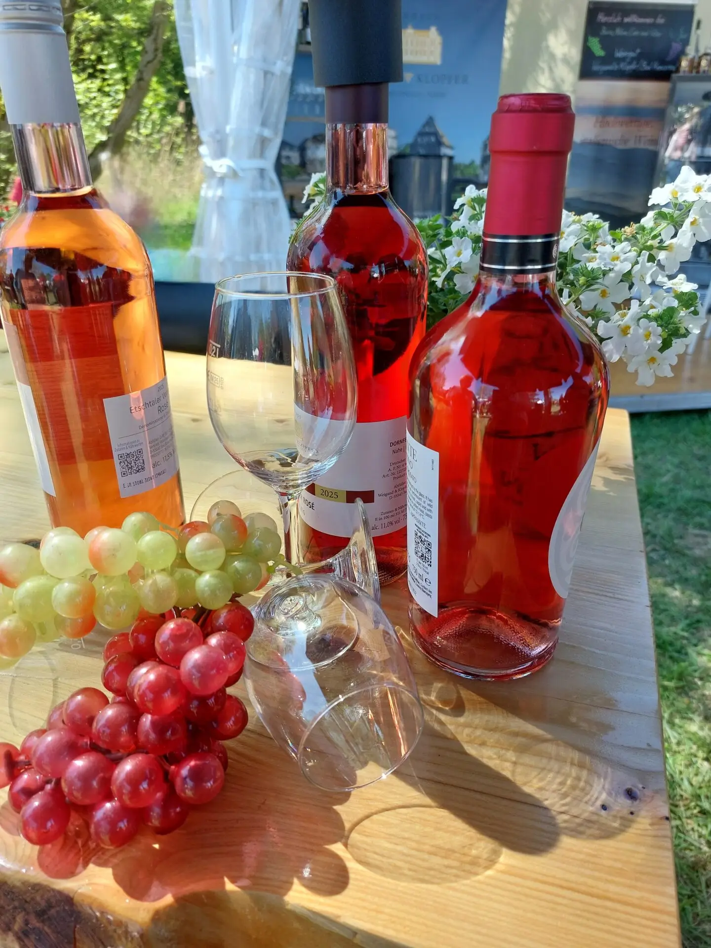Drei Flaschen Roséwein aus dem Weinland, zwei Weingläser, dekorative grüne und rote Weintrauben und weiße Blumen auf einem Holztisch im Freien - perfekt für Ihre Think Pink Feier am 18.09.2026 mit Grünzeug und Bannern im Hintergrund.
